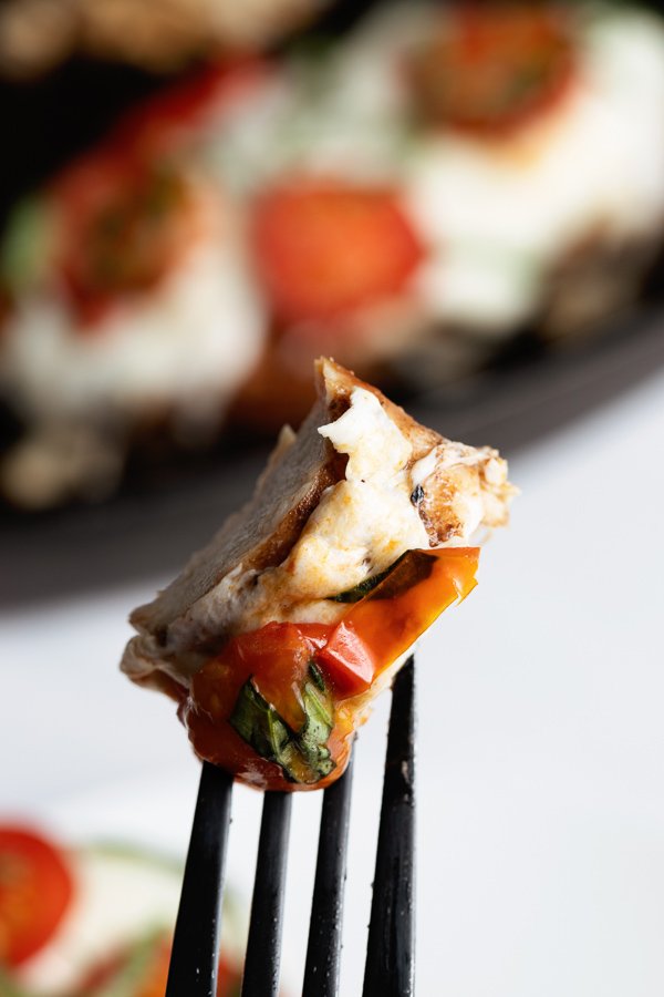 A fork holds a bite meat with layers of melted cheese, sliced tomatoes, and fresh green herbs against a blurred background of a similar dish.