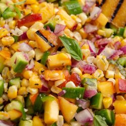 A vibrant salad with diced cucumbers, sweet corn, red onions, and peaches, garnished with fresh basil leaves and charred lemon wedges.