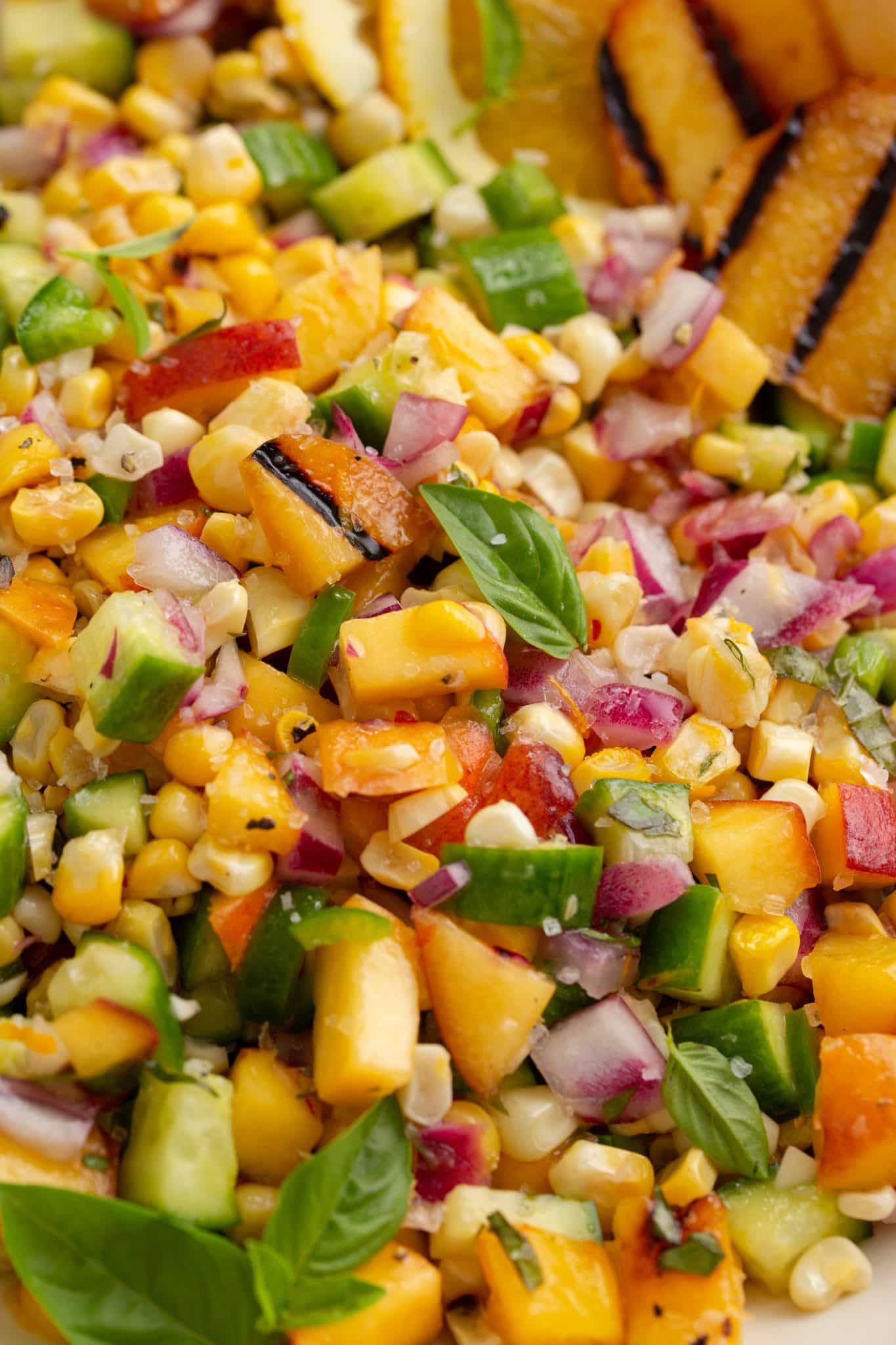A vibrant salad with diced cucumbers, sweet corn, red onions, and peaches, garnished with fresh basil leaves and charred lemon wedges.
