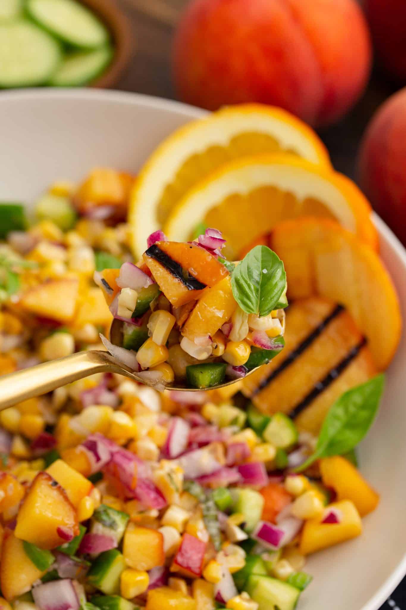 A spoonful of salad with grilled peaches, corn, red onions, cucumbers, and basil shown over a bowl of the same salad. Slices of orange and whole peaches are in the background.