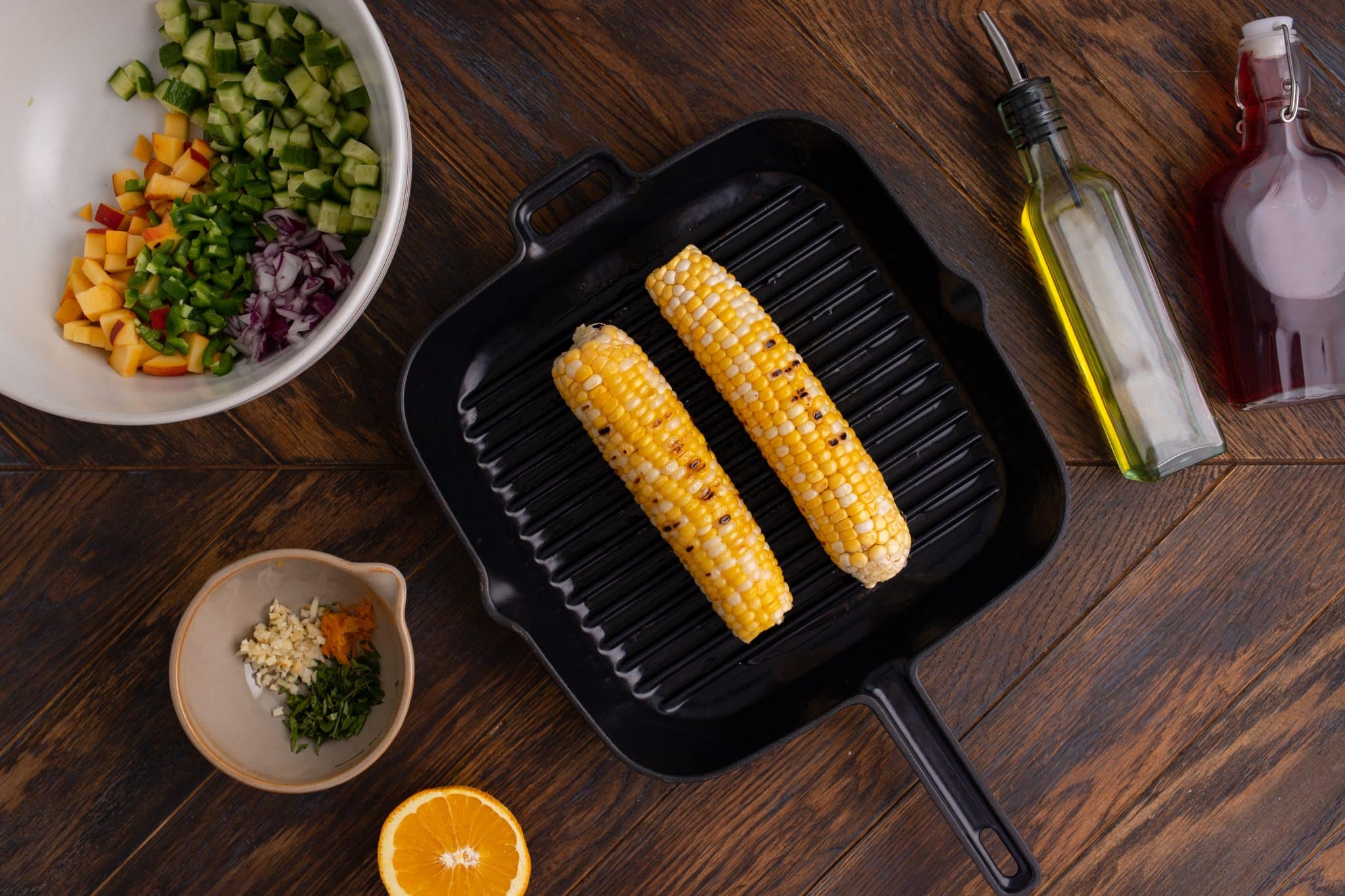 Two grilled corn cobs in a grill pan surrounded by a bowl of chopped vegetables, a small bowl of chopped garlic and herbs, a halved orange, a bottle of olive oil, and a bottle of sauce on a wooden table.