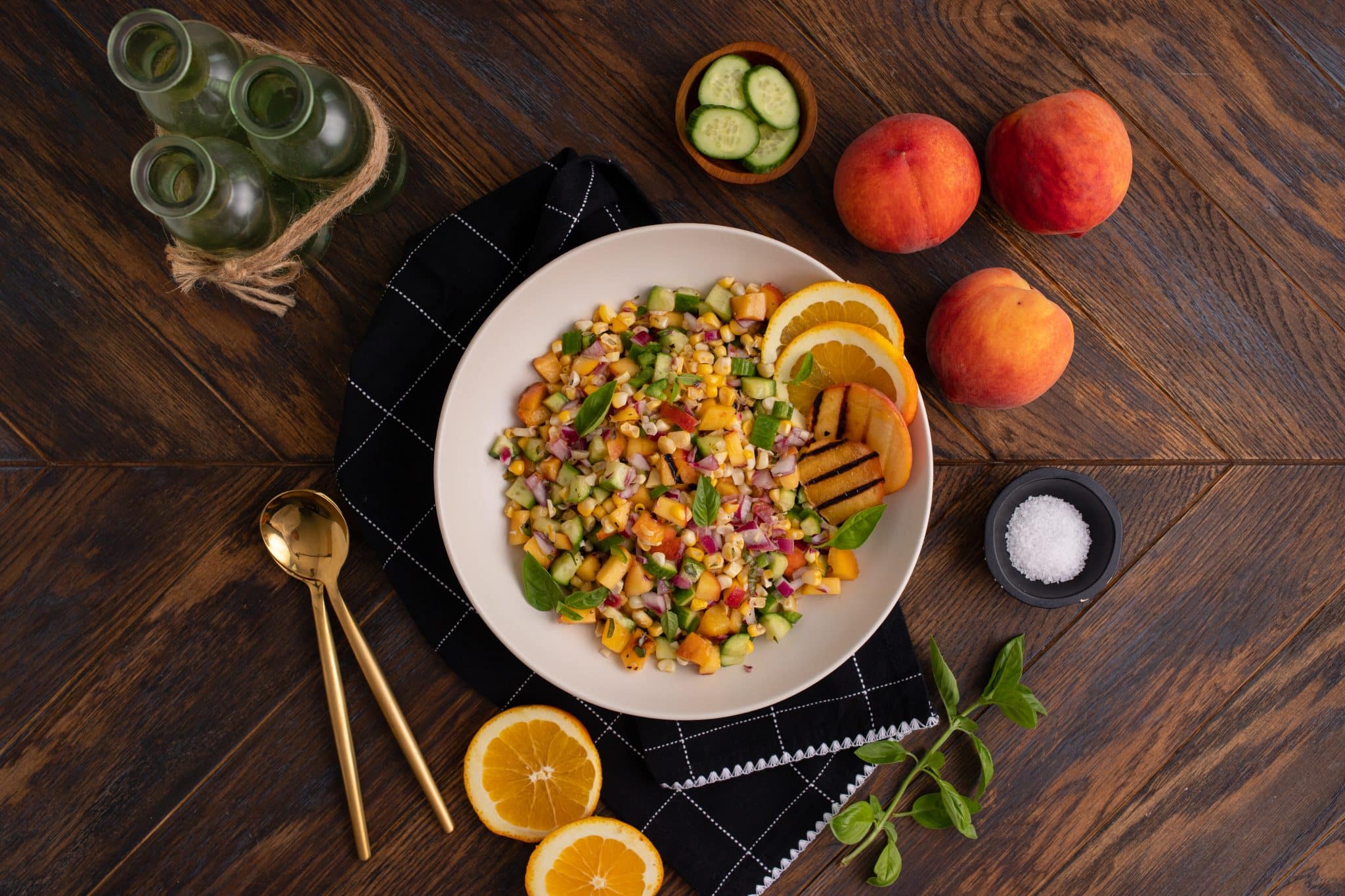 A white bowl filled with a mixed salad featuring diced vegetables, beans, and fresh peaches, accompanied by sliced oranges, cucumbers, whole peaches, gold cutlery, and a black napkin on a wooden table.