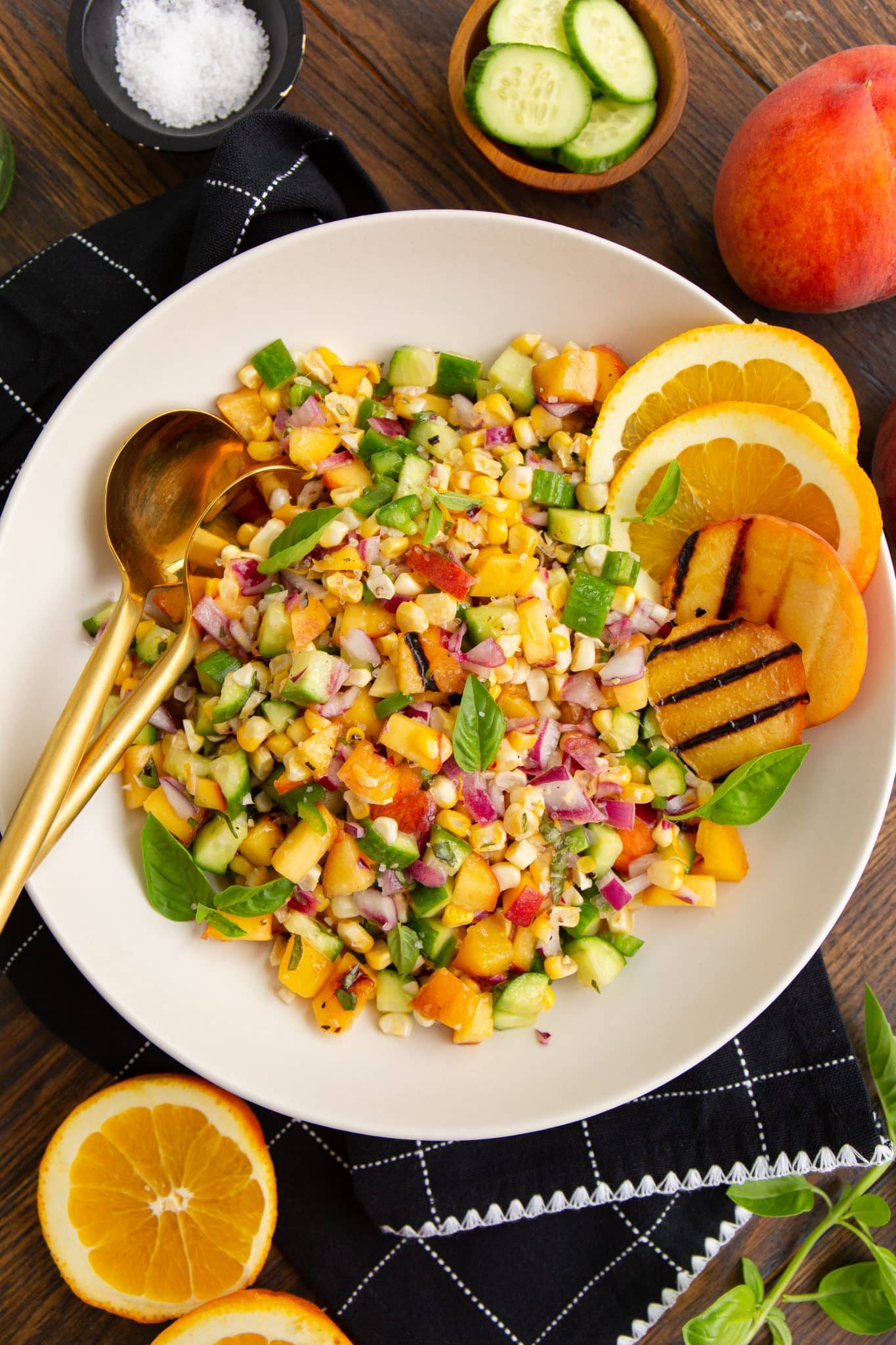 A colorful salad with chopped vegetables and fruits in a white bowl, garnished with basil leaves. Sliced oranges and grilled fruit are on the side, with metal utensils placed on the rim of the bowl.