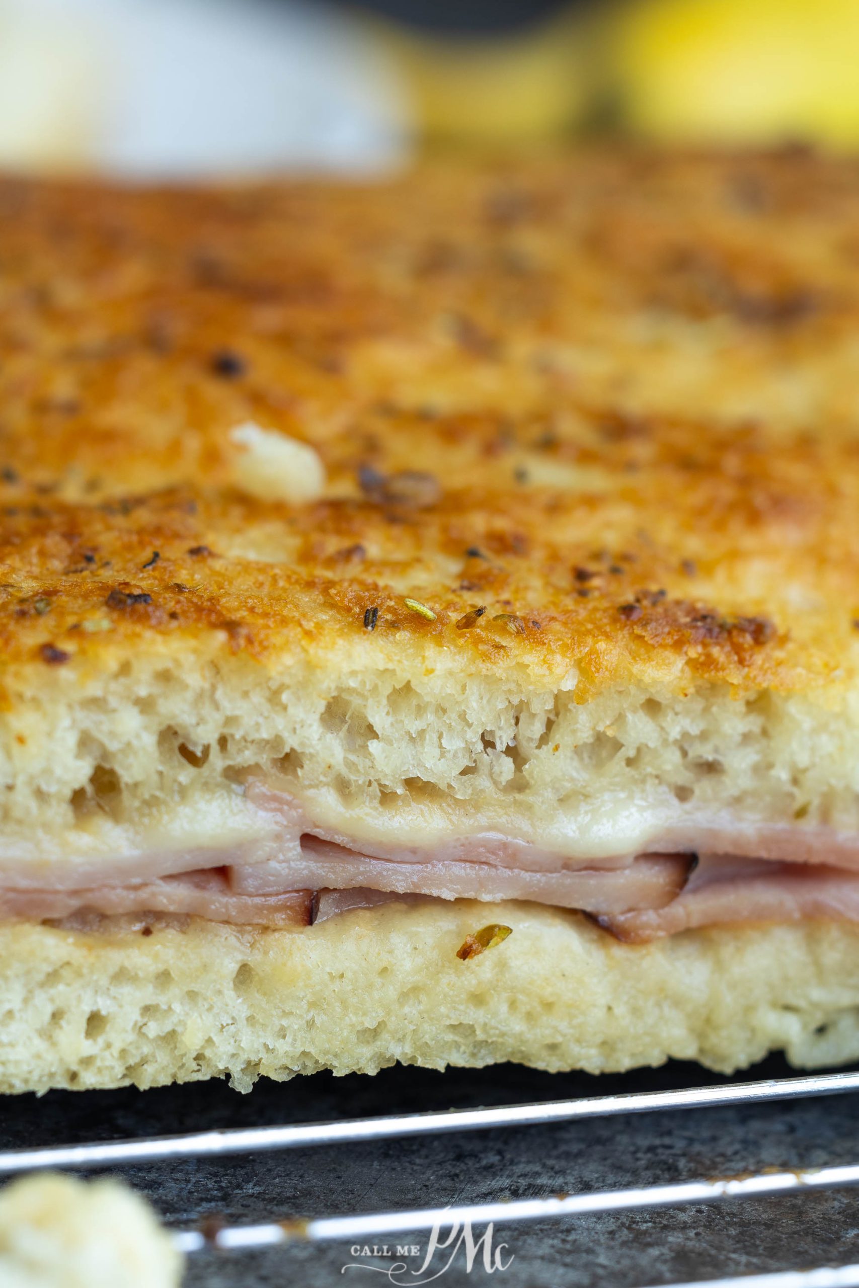 A close-up image of a sandwich with layers of bread, melted cheese, and sliced ham, showcasing a golden-brown crust with herbs.