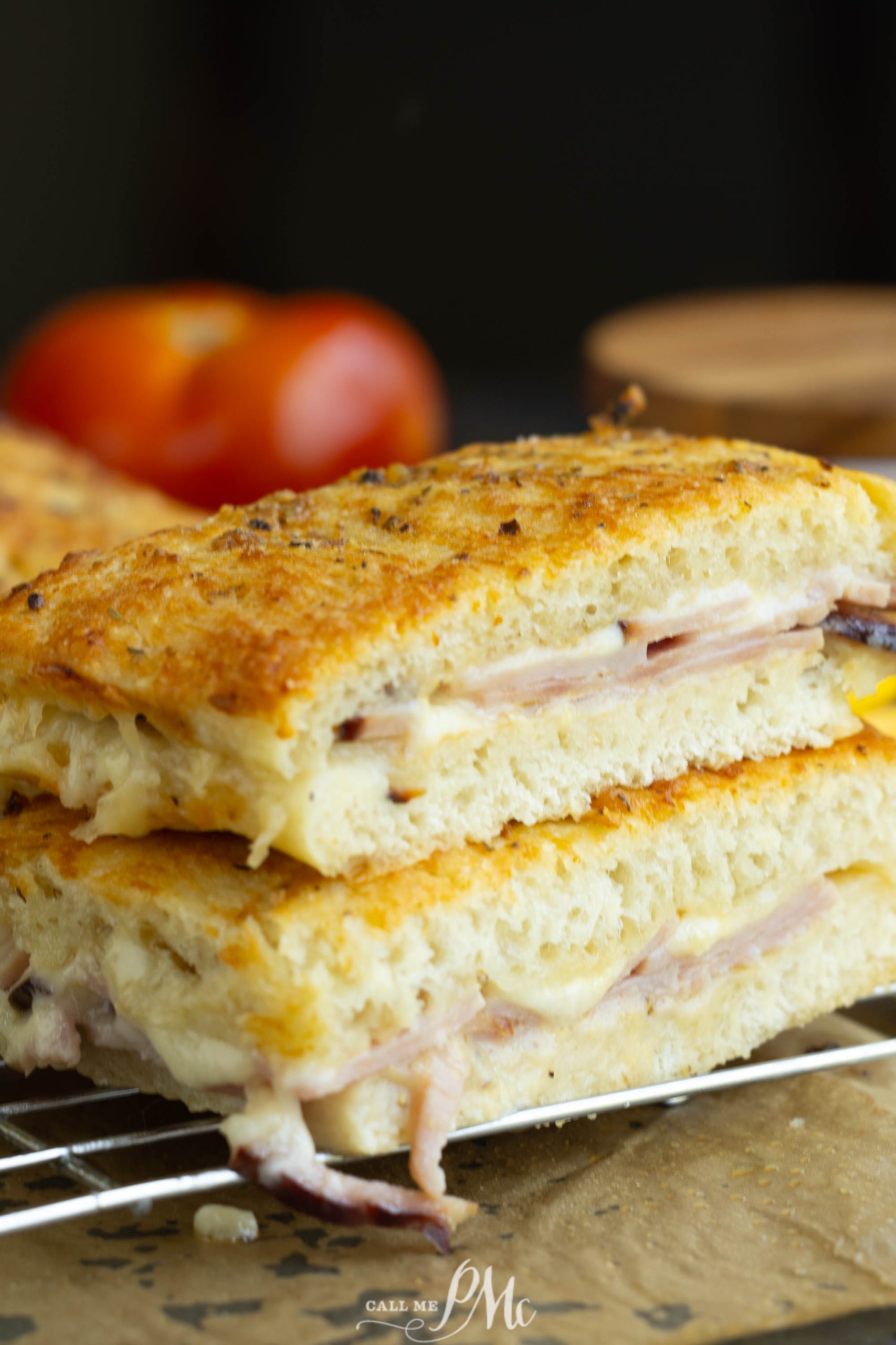 A close-up of a toasted sandwich with ham and melted cheese on a wire rack. There is a partially visible red tomato in the background.