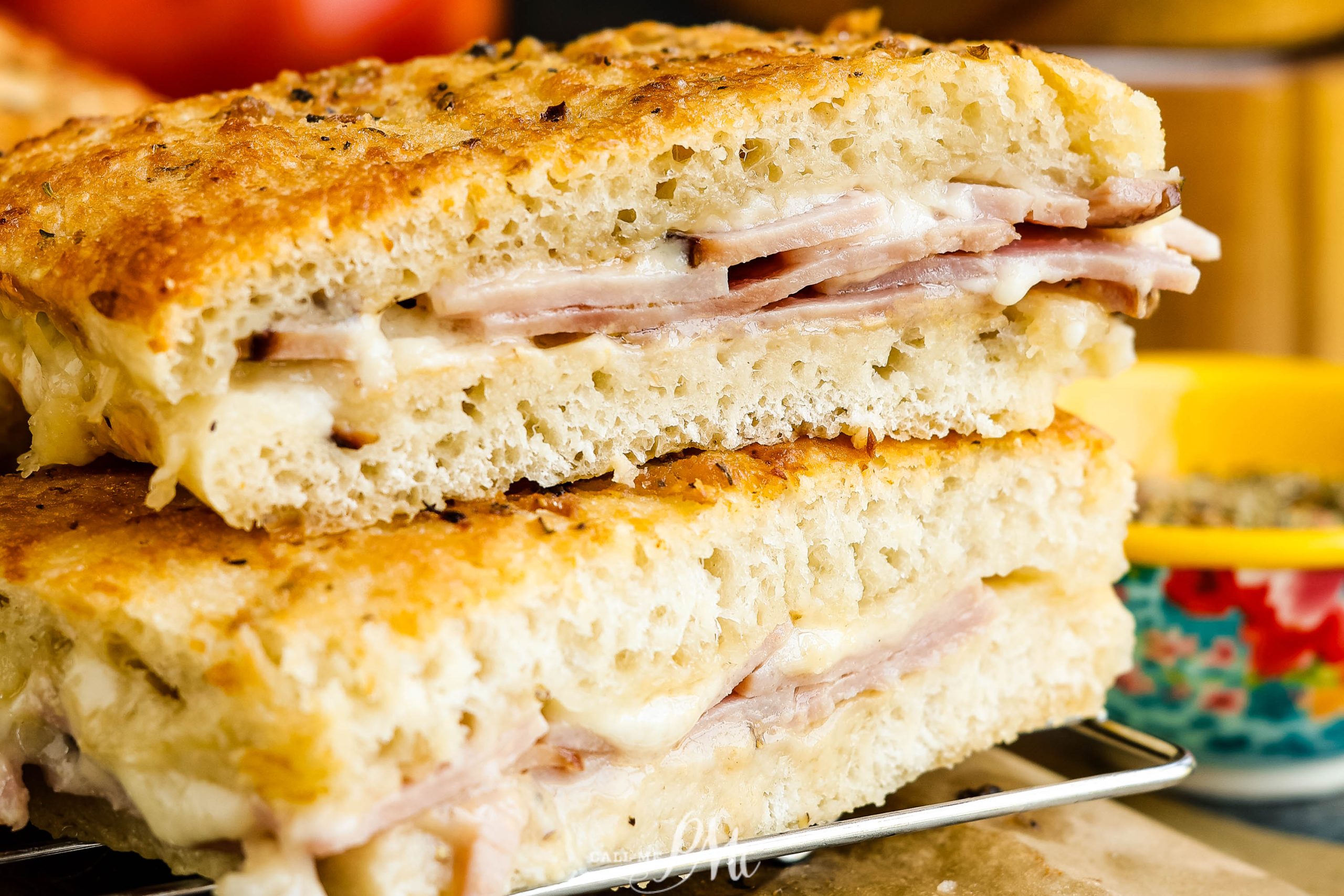 Close-up of two halves of a sandwich made with thick focaccia bread, filled with layers of ham and melted cheese. The sandwich is placed on a wire rack.