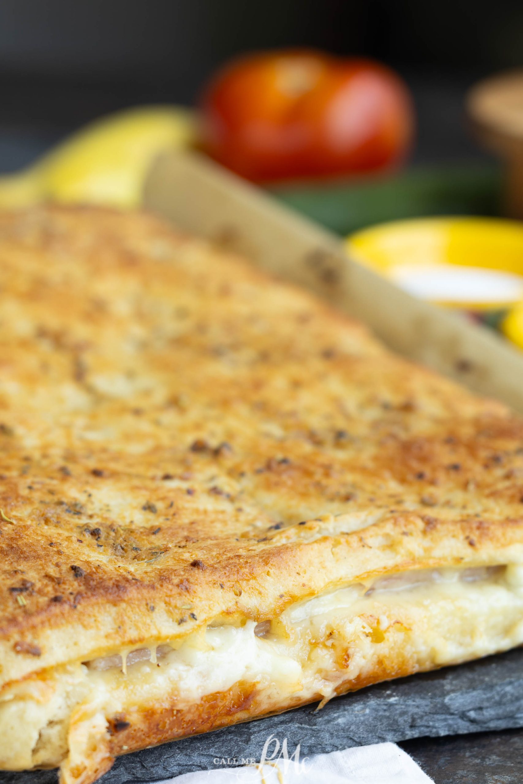 A close-up of a golden-brown rectangular baked dish filled with melted cheese. A tomato and lemon are blurred in the background.