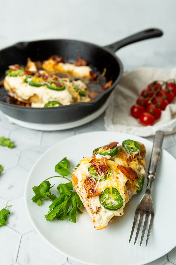 A plate with a serving of stuffed chicken breast topped with cheese, bacon, and jalapeño slices. Fork on the plate, fresh tomatoes and cilantro on the side, remaining chicken in a skillet in the background.