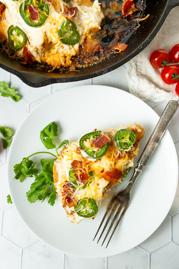 A plate with a serving of stuffed chicken breast topped with cheese, bacon, and jalapeño slices. Fork on the plate, fresh tomatoes and cilantro on the side, remaining chicken in a skillet in the background.