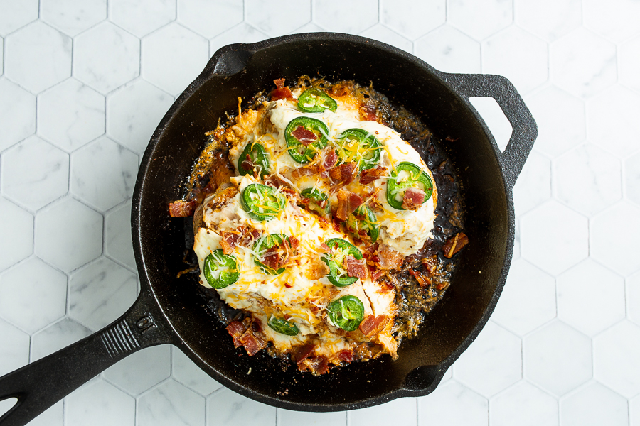 A cast iron skillet containing jalapeno popper chicken topped with melted cheese, bacon bits, and sliced jalapeños on a hexagon-tile background.