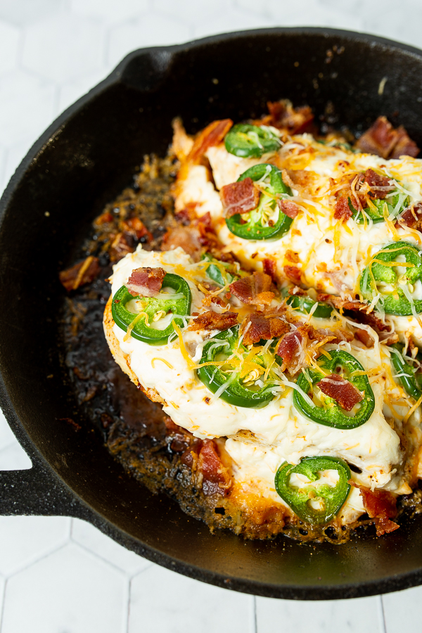 A cast iron skillet with cooked chicken breasts topped with melted cheese, jalapeño slices, and bacon bits.