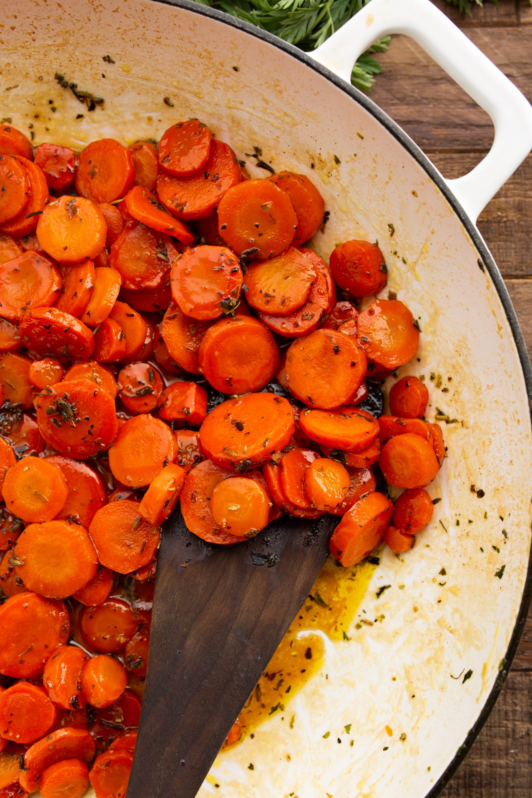 A pan of glazed, cooked carrot slices with a wooden spatula, seasoned with herbs and oil.