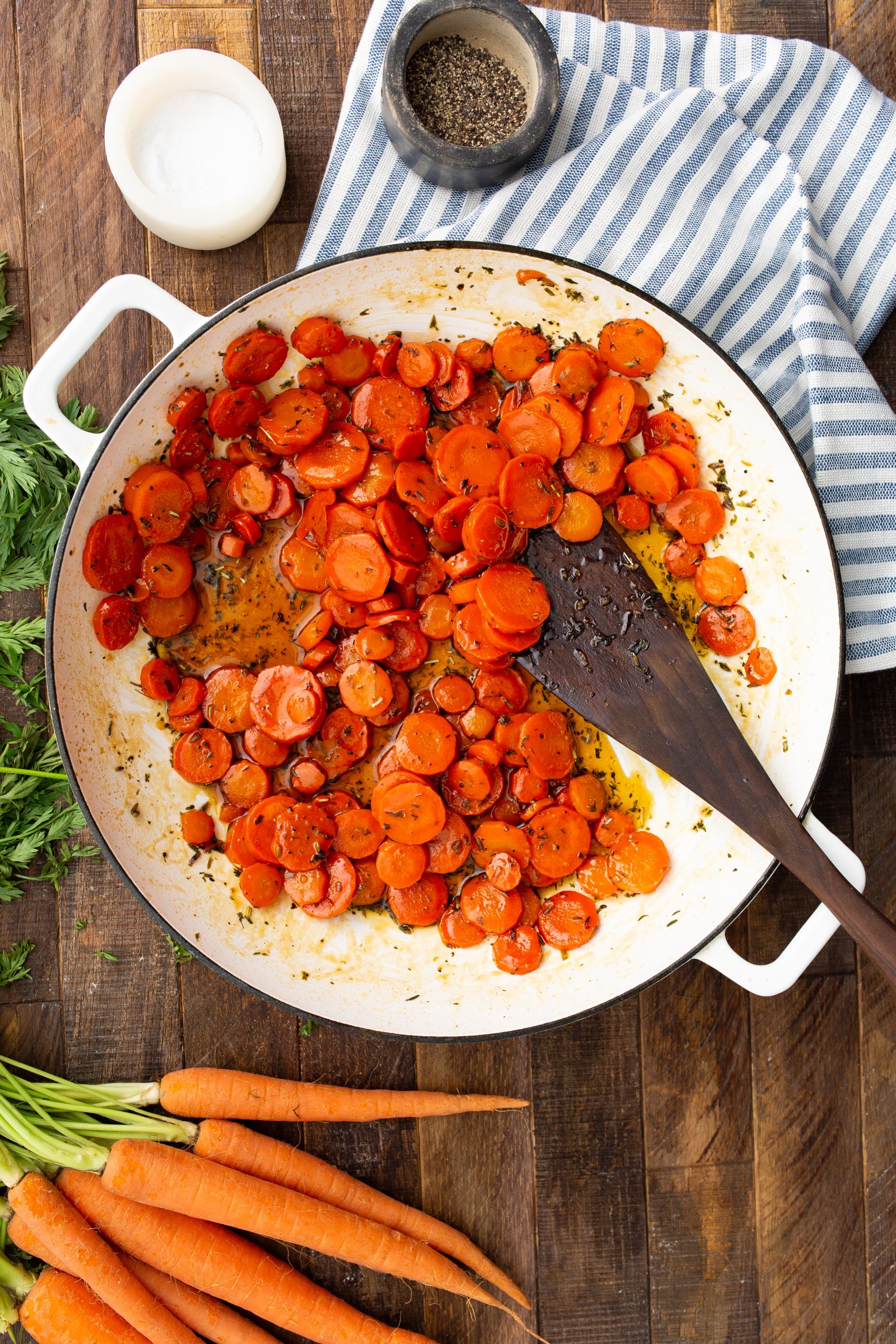 A white pan filled with Maple Cider Glazed Carrots  is placed on a wooden surface, accompanied by a wooden spatula, fresh carrots, a blue striped cloth, and small containers of salt and pepper.