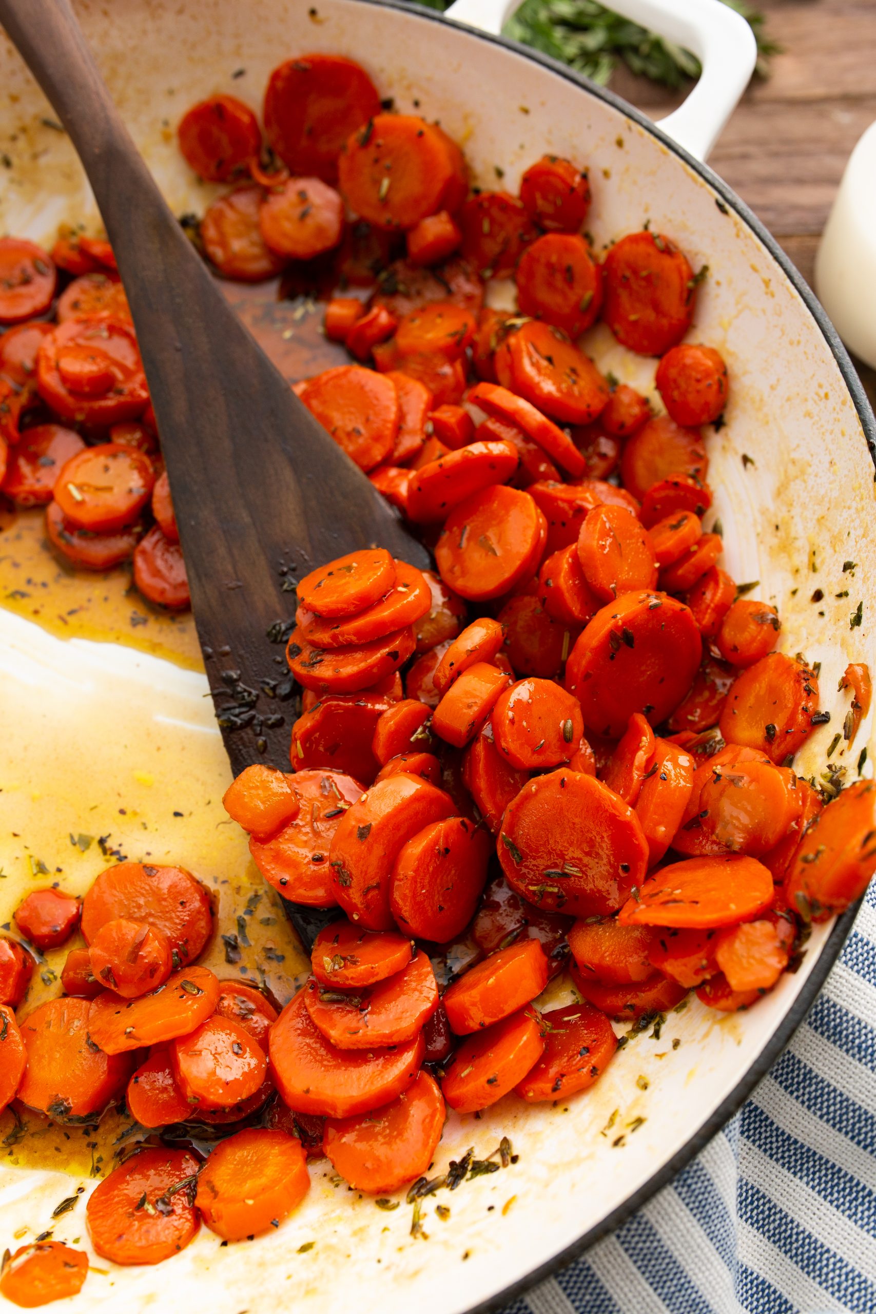 A close-up of sliced, cooked carrots seasoned with herbs in a white pan, being stirred with a wooden spoon.