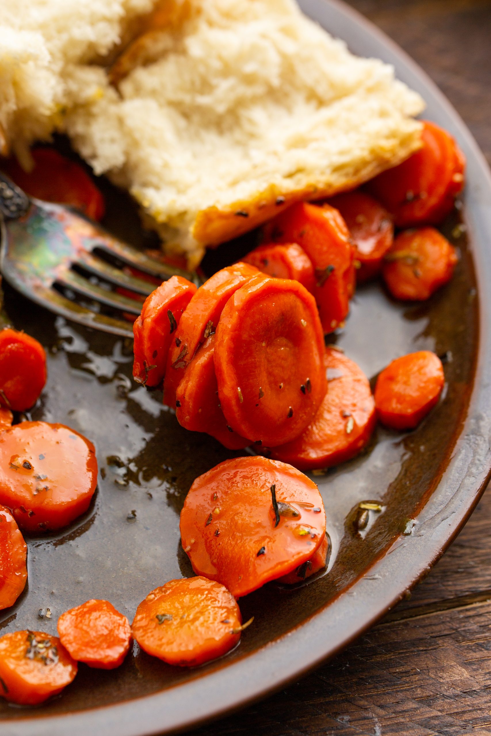 A close-up of a plate with sliced cooked carrots, seasoned with herbs, next to a piece of bread. A fork rests on the plate.