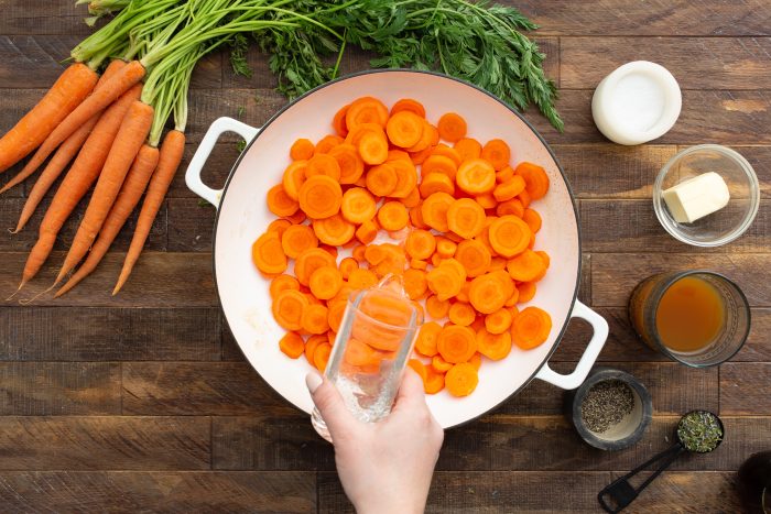 A hand is pouring water over sliced carrots in a pan with whole carrots, salt, butter, broth, pepper, and a measuring spoon arranged on a wooden surface.