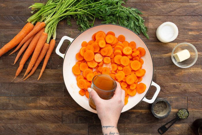 A hand pouring liquid into a pan filled with sliced carrots, surrounded by fresh carrots, salt, butter, and pepper on a wooden table.