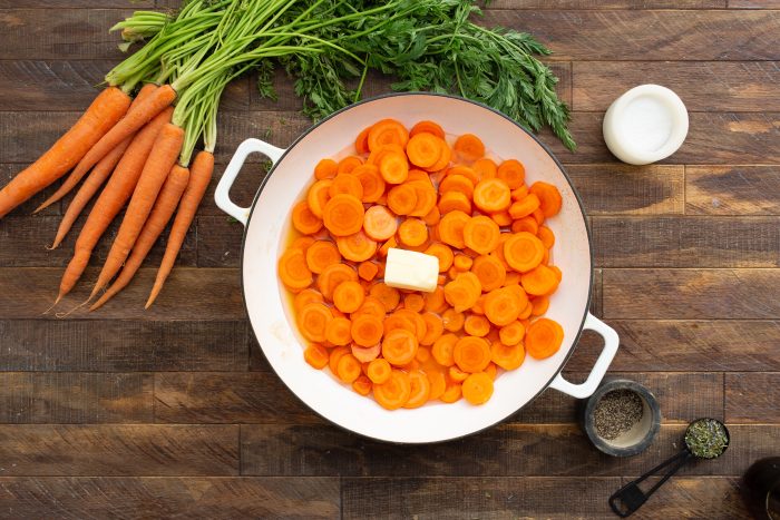 Sliced carrots and a pat of butter in a white pan, surrounded by fresh whole carrots, a bowl of salt, and a bowl of herbs on a wooden surface.