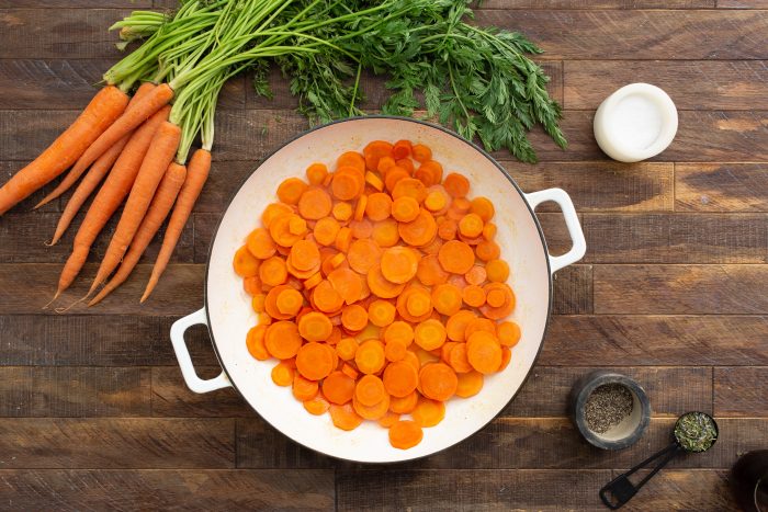 Sliced carrots in a white pan on a wooden surface, with fresh whole carrots, a small bowl of black pepper, chopped herbs, and a container of salt nearby.