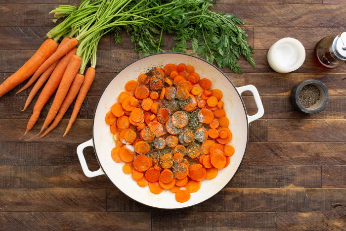 A white pan filled with sMaple Cider Glazed Carrots  with herbs, placed on a wooden surface next to whole carrots, a small bowl of salt, a pepper grinder, and a bottle of oil.