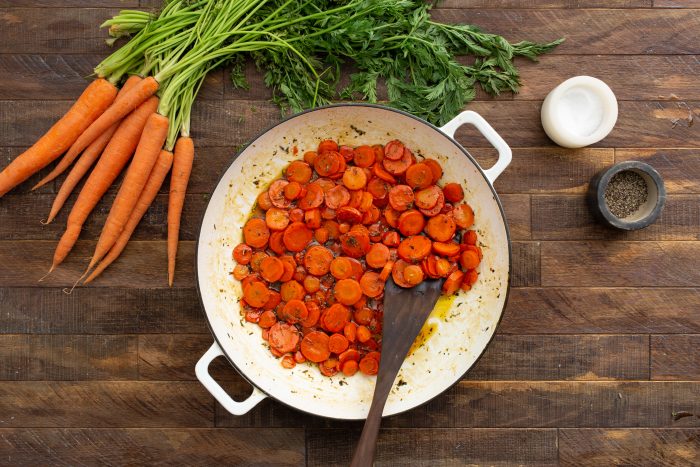 A white pan with Maple Cider Glazed Carrots  in a red sauce on a wooden table, surrounded by fresh carrots and two small bowls, one containing pepper and the other empty.