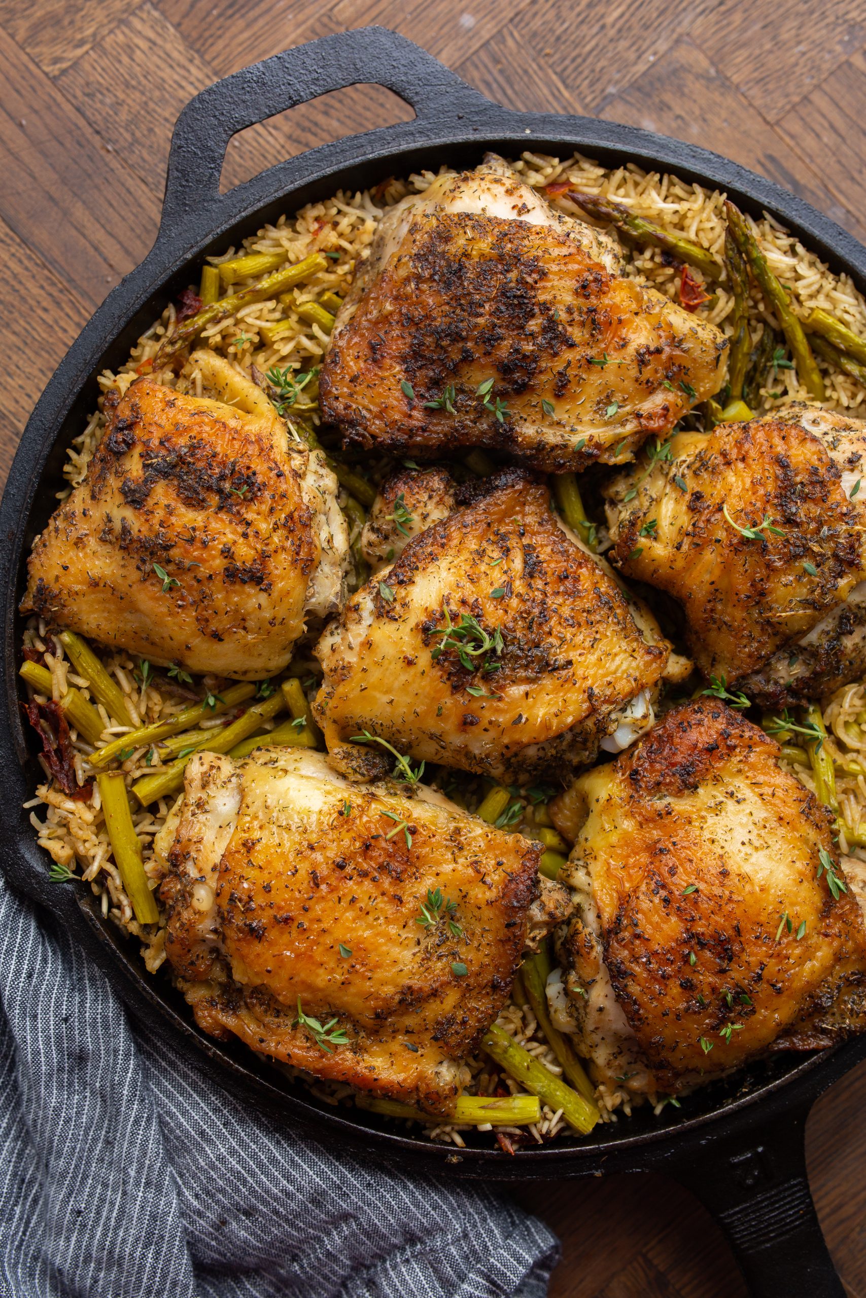 A cast-iron skillet filled with seasoned, roasted chicken thighs on a bed of rice mixed with green vegetables, garnished with thyme and lemon wedges.