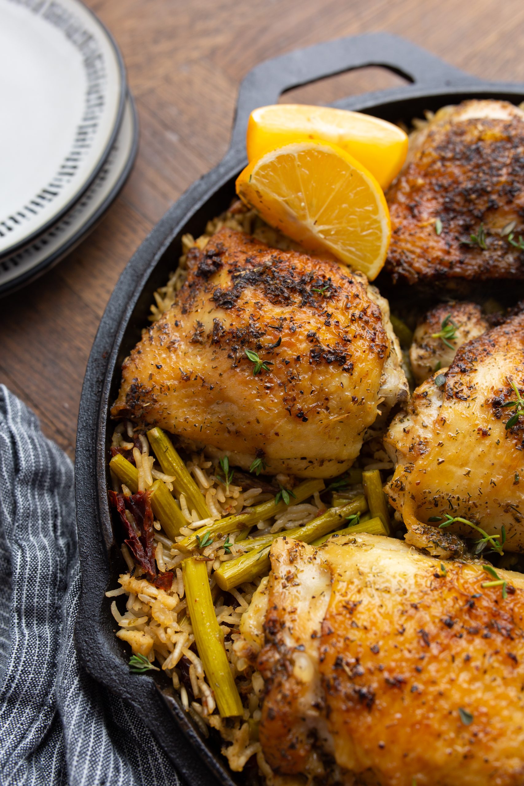 A cast-iron skillet filled with seasoned, roasted chicken thighs on a bed of rice mixed with green vegetables, garnished with thyme and lemon wedges.