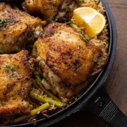 A cast-iron skillet filled with seasoned, roasted chicken thighs on a bed of rice mixed with green vegetables, garnished with thyme and lemon wedges.