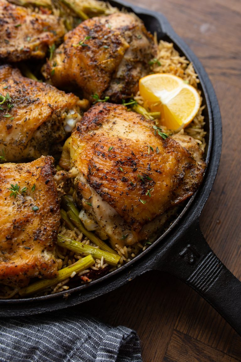 A cast-iron skillet filled with seasoned, roasted chicken thighs on a bed of rice mixed with green vegetables, garnished with thyme and lemon wedges.