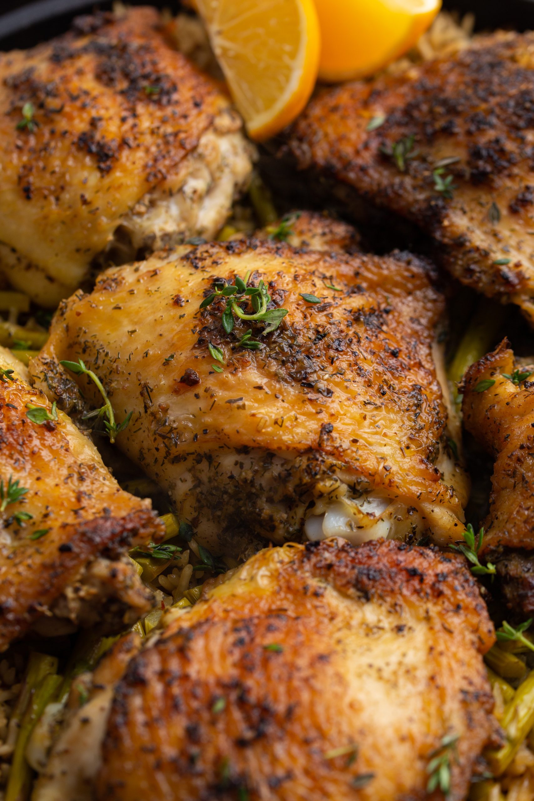 A cast-iron skillet filled with seasoned, roasted chicken thighs on a bed of rice mixed with green vegetables, garnished with thyme and lemon wedges.