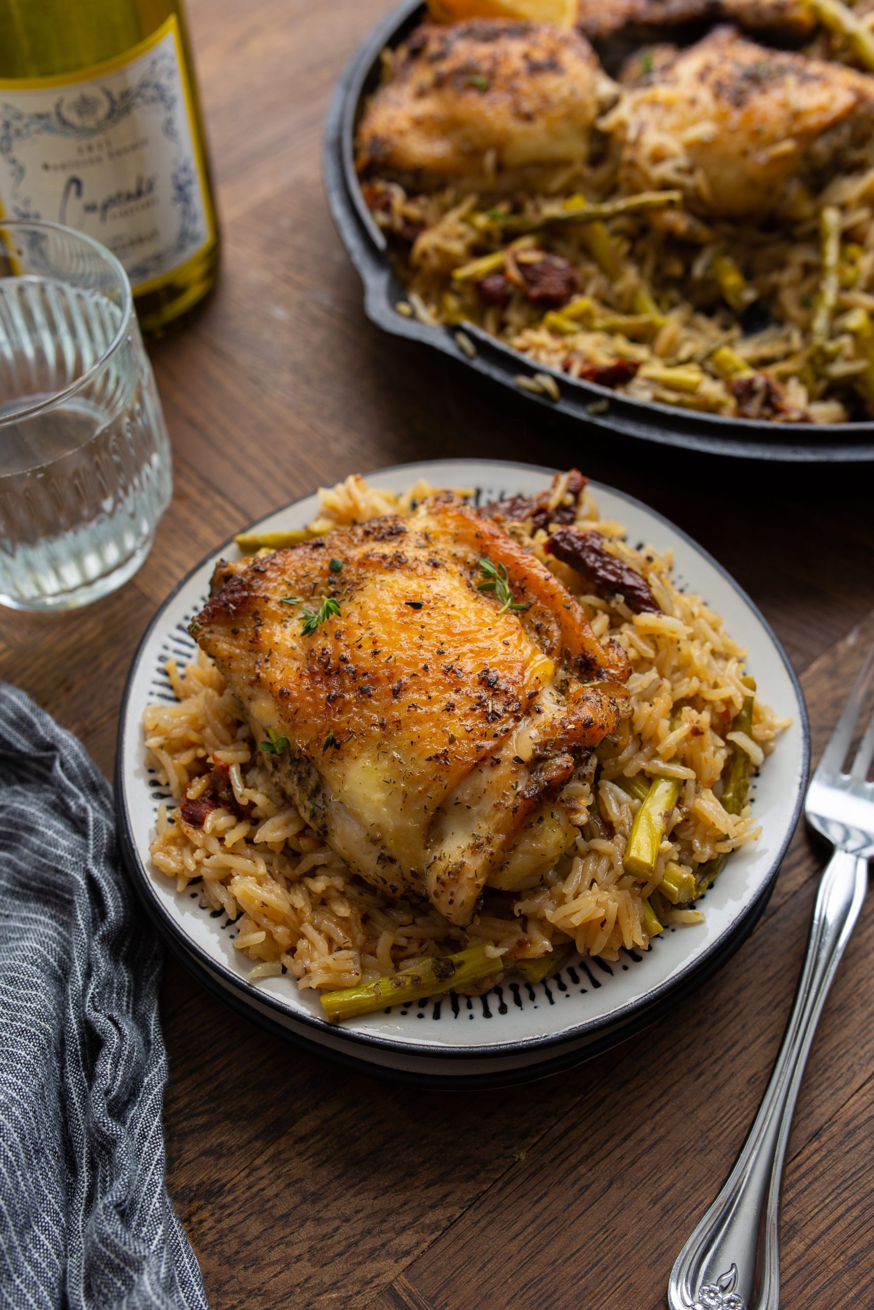 A cast-iron skillet filled with seasoned, roasted chicken thighs on a bed of rice mixed with green vegetables, garnished with thyme and lemon wedges.