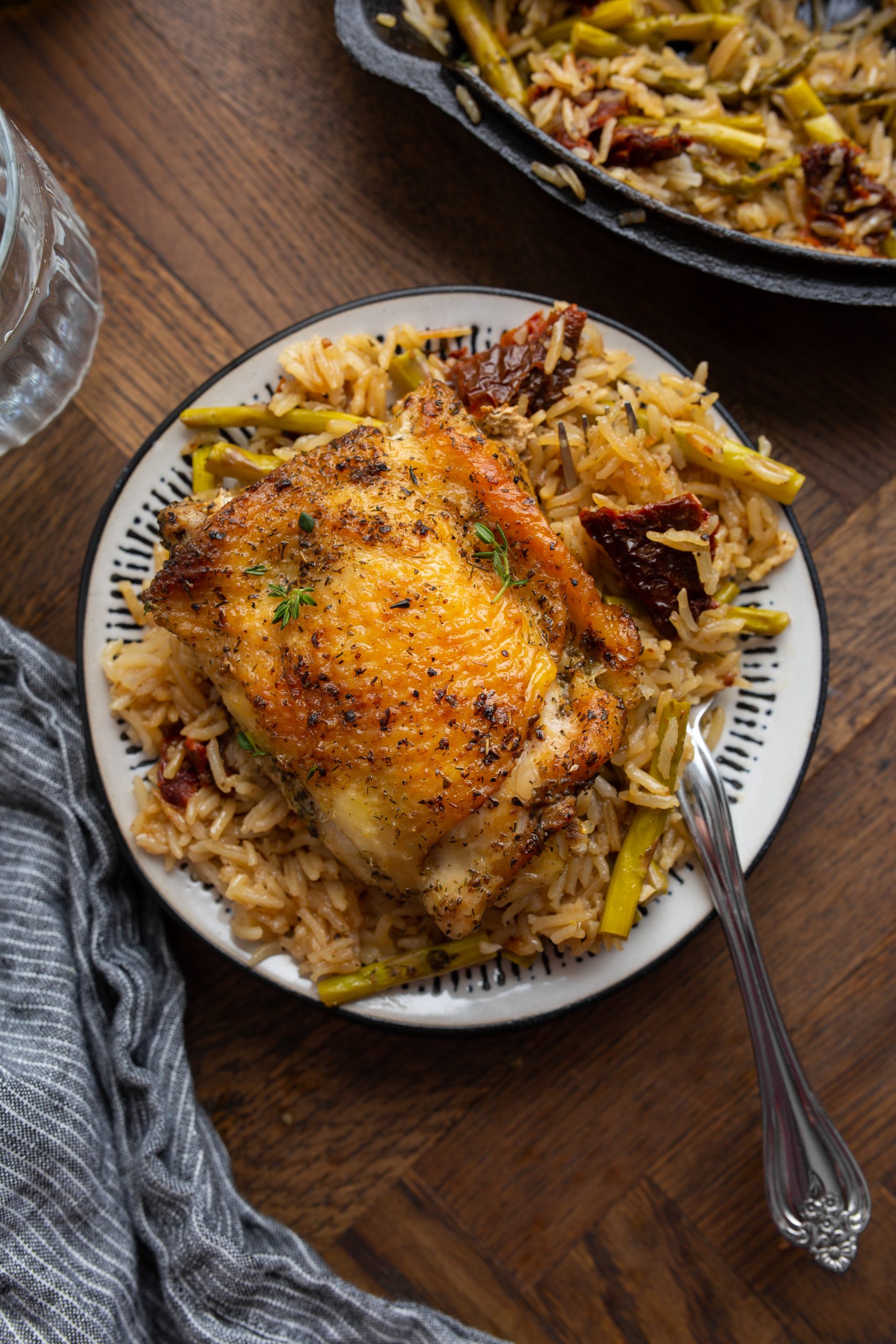 A cast-iron skillet filled with seasoned, roasted chicken thighs on a bed of rice mixed with green vegetables, garnished with thyme and lemon wedges.