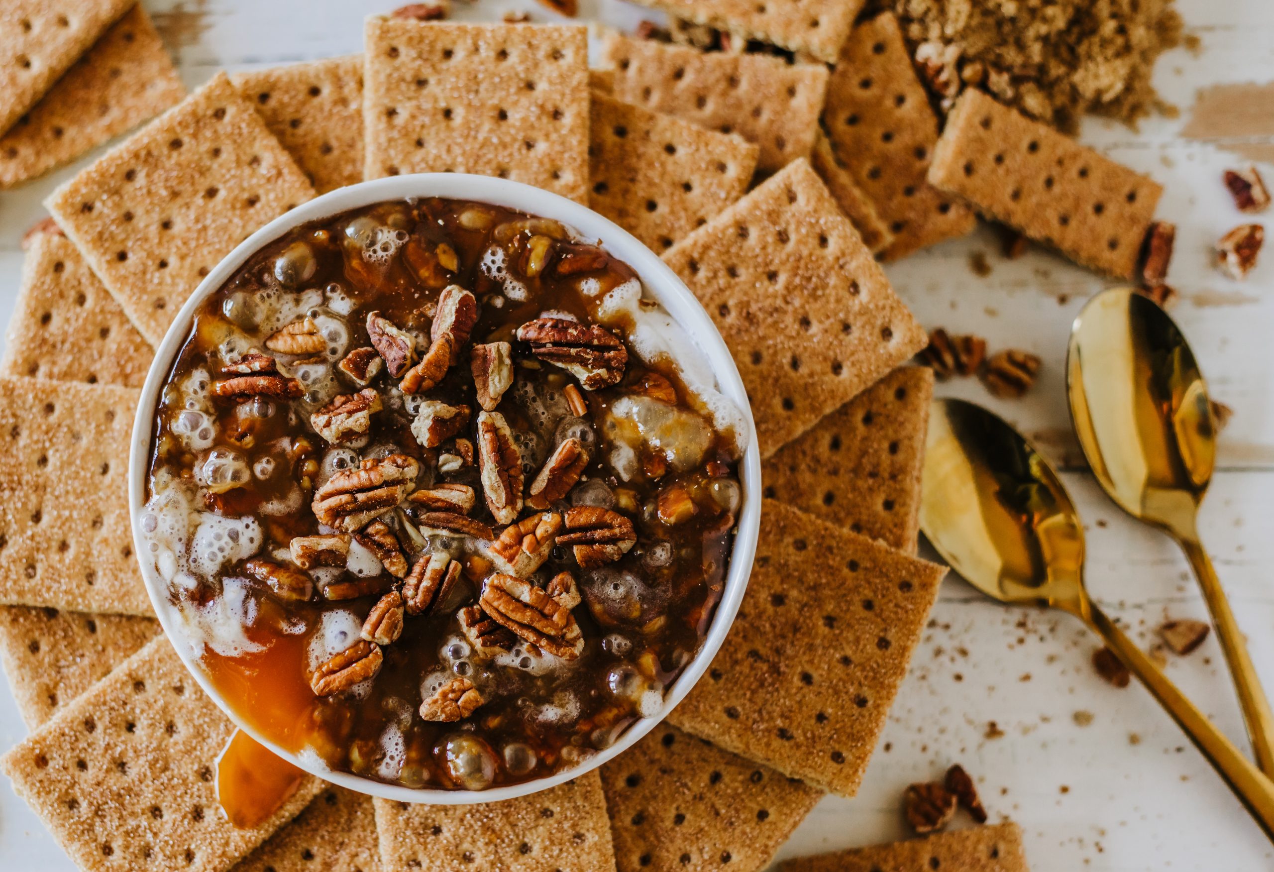 A bowl filled with pecan-topped dessert surrounded by graham crackers. Two gold spoons are placed to the right.