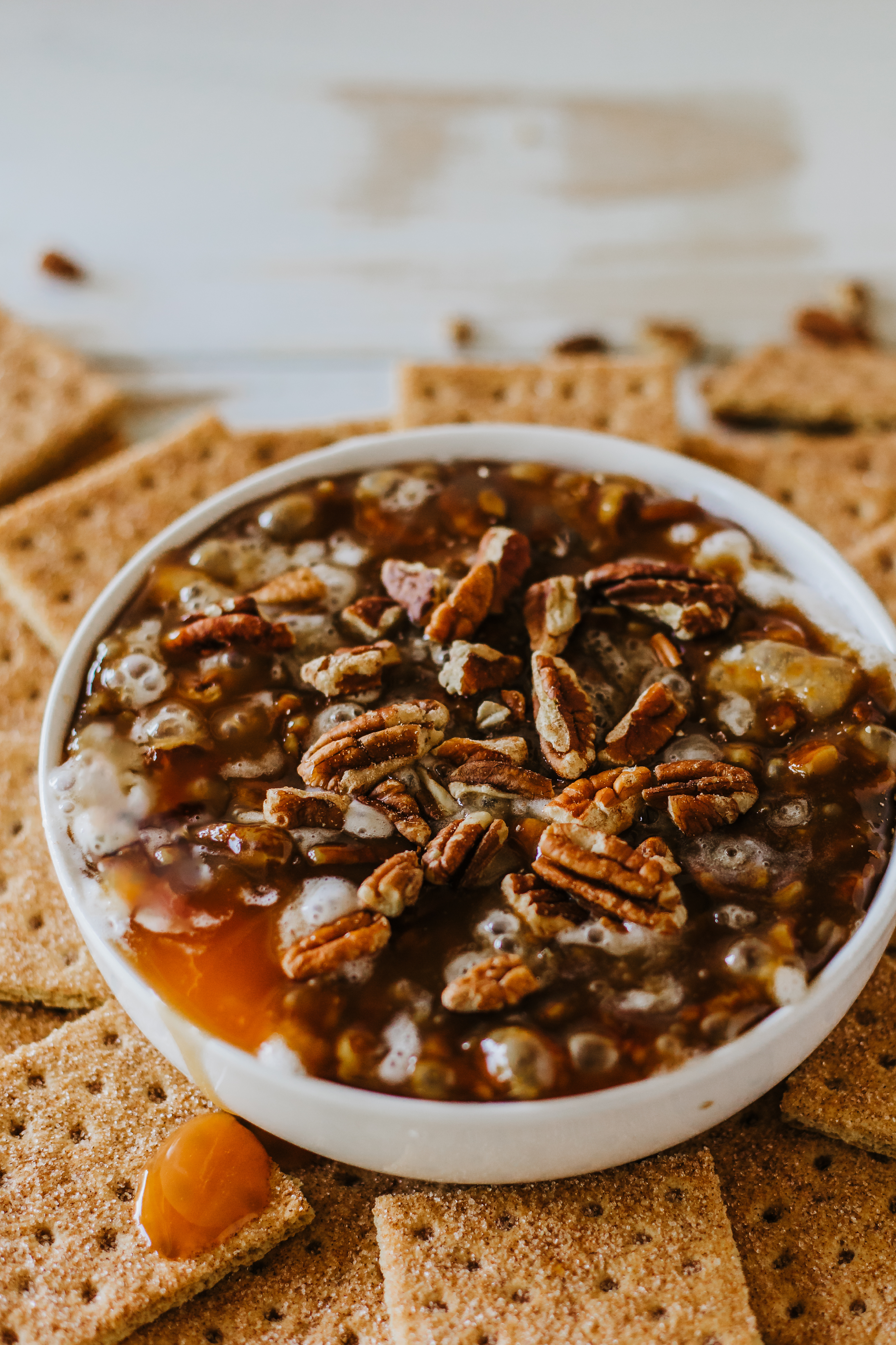 A bowl filled with pecan-topped dessert surrounded by graham crackers. Two gold spoons are placed to the right.