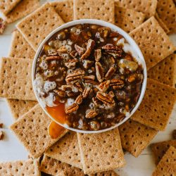 A bowl filled with pecan-topped dessert surrounded by graham crackers. Two gold spoons are placed to the right.