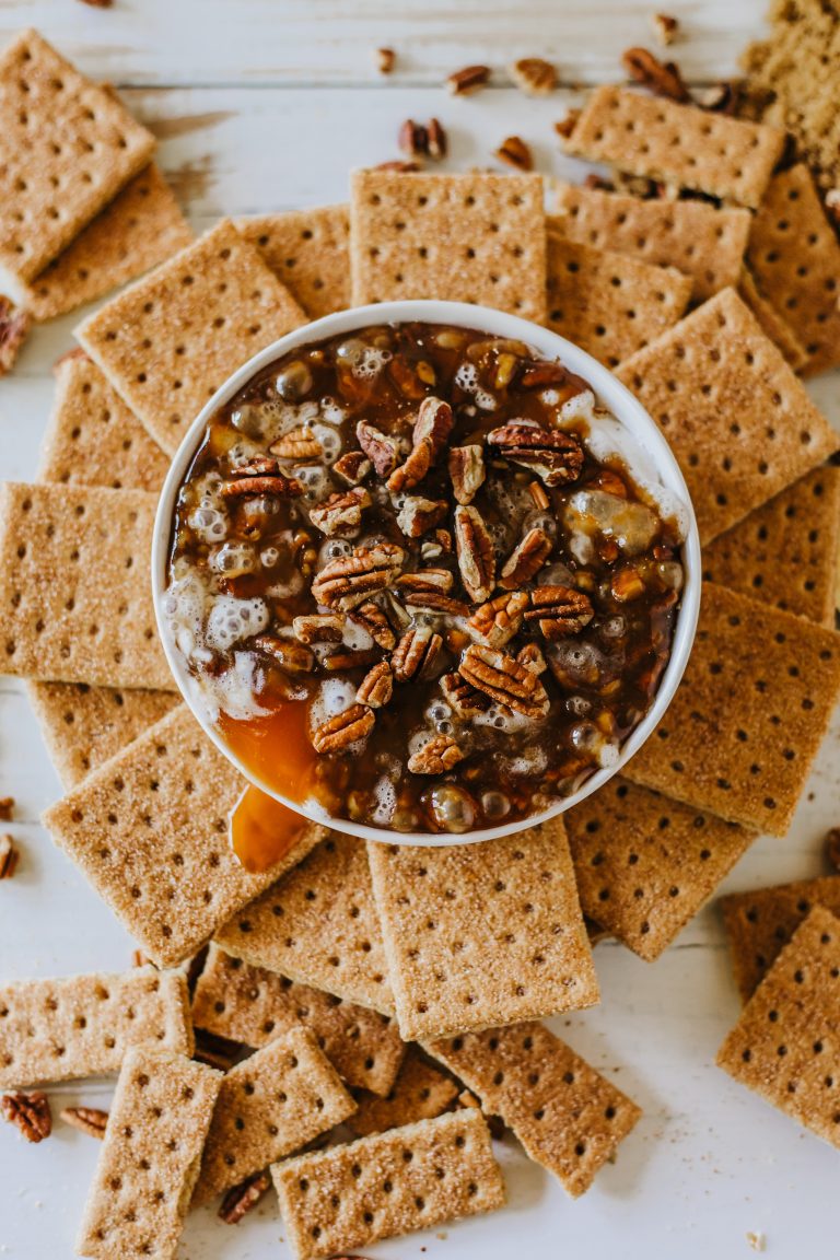 A bowl filled with pecan-topped dessert surrounded by graham crackers. Two gold spoons are placed to the right.