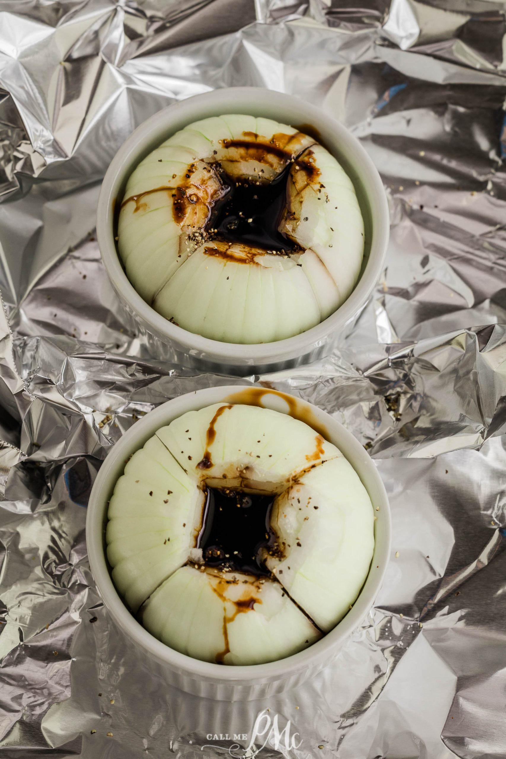 Two partially cut onions sit in white ramekins, filled with balsamic glaze and seasoning, placed on a crinkled aluminum foil background.