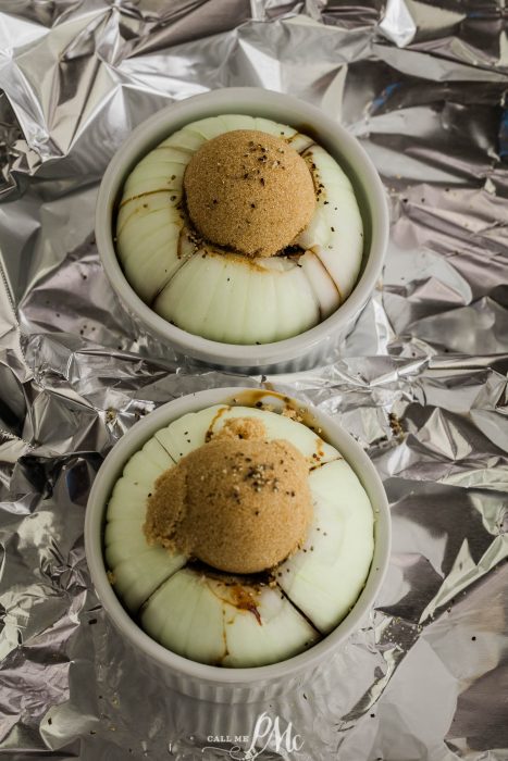 Two onions in small white ramekins, topped with round portions of brown sugar and seasoning, are placed on crumpled aluminum foil.