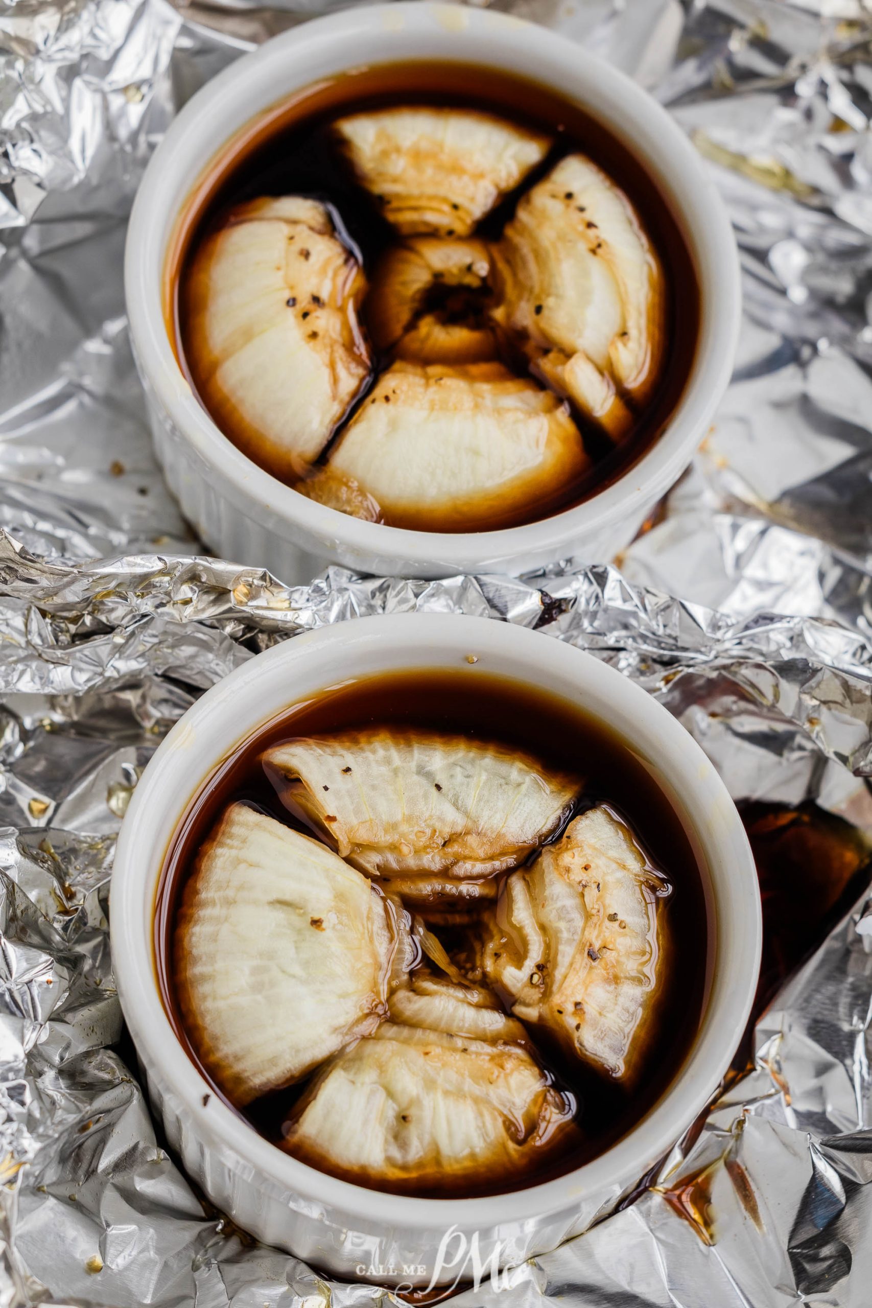 Two white ramekins containing sliced onions in a dark liquid, placed on a surface covered with aluminum foil.