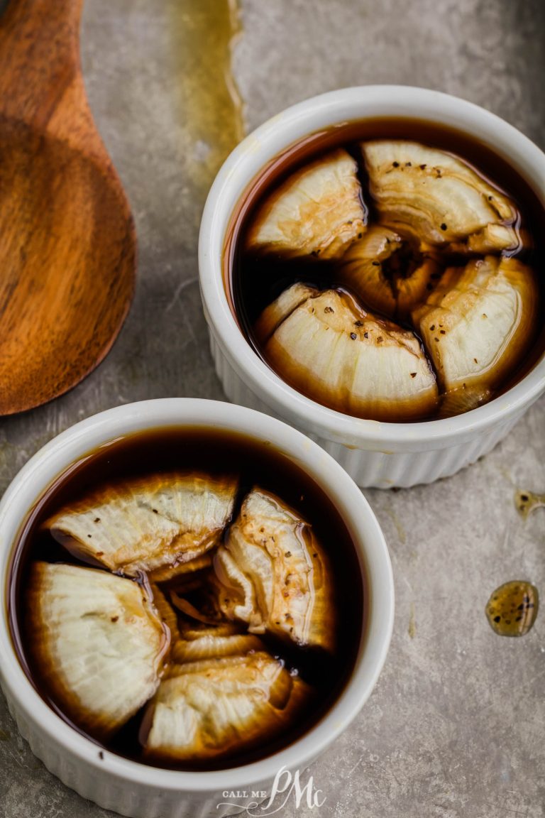 Two white ramekins filled with brown liquid and sliced onions, placed on a gray surface. A wooden spoon lies to the side.