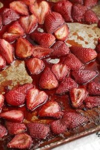 Halved strawberries with a roasted appearance are spread out on a metal baking sheet.