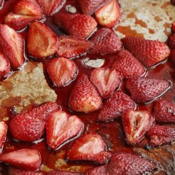 Halved strawberries with a roasted appearance are spread out on a metal baking sheet.