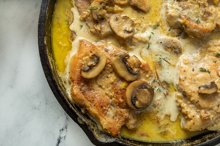 A close-up of a skillet dish featuring seared pork chops topped with mushrooms and simmering in a creamy sauce.