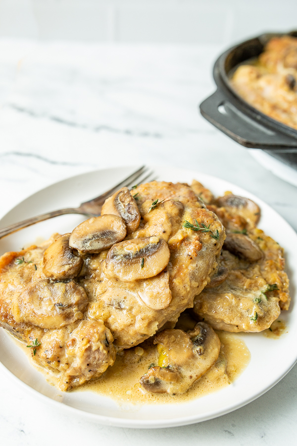 A plate with creamy pork chops topped with sliced mushrooms and a fork on the side. A pan with more pork chops is partially visible in the background.