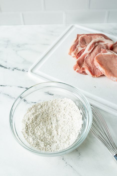 A glass bowl of flour mixture, a whisk, and raw pork chops ready to be turned into smothered pork chops on a white cutting board on a marble countertop.