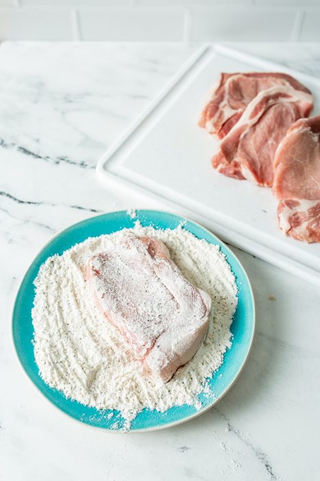 A piece of raw pork is coated in flour on a blue plate. Other pieces of raw pork are on a white cutting board in the background.