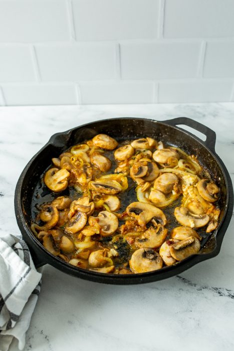 A cast-iron skillet filled with sautéed mushrooms and onions placed on a white marble surface. A checkered cloth is partially visible on the left side.