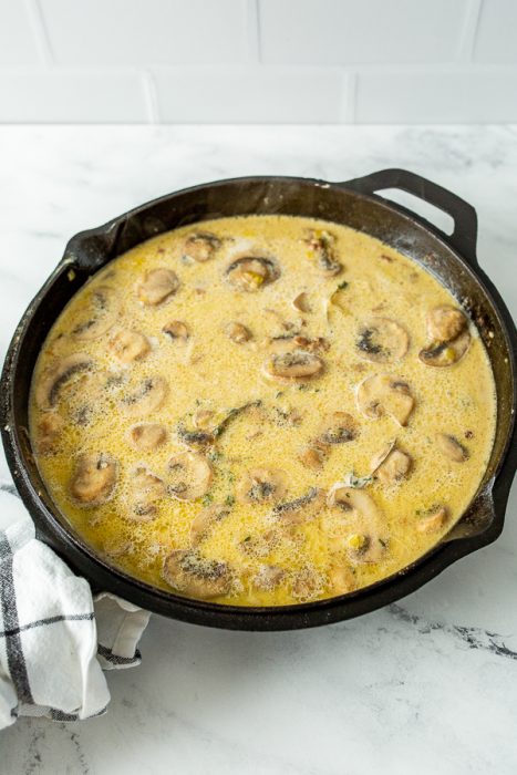 A black skillet filled with a creamy mushroom sauce, featuring sliced mushrooms in a light-colored liquid, sitting on a white marble surface next to a folded black and white checkered kitchen towel.