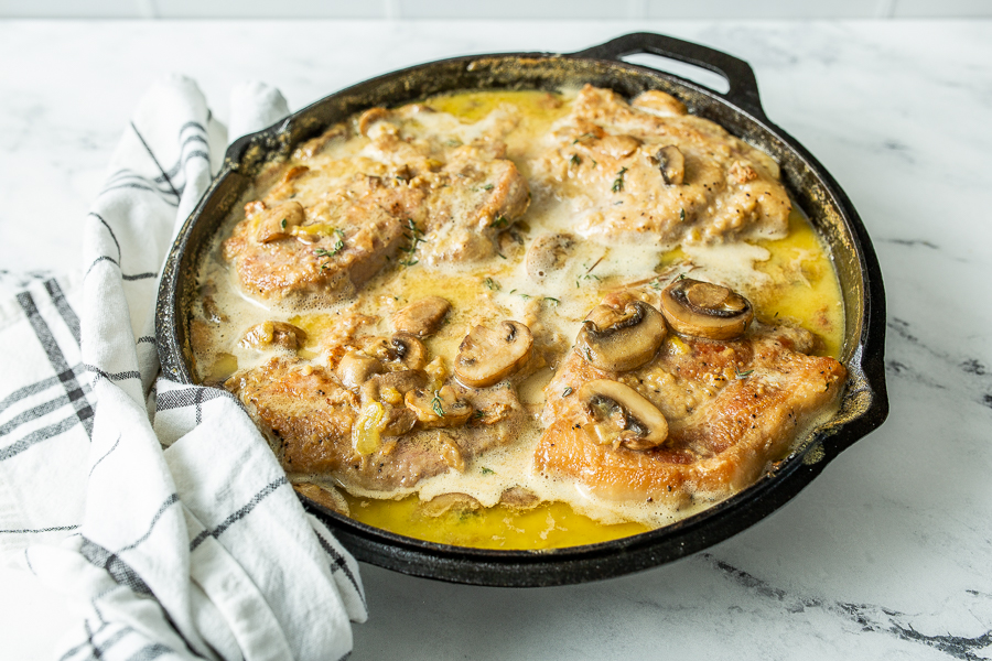 A cast iron skillet filled with cooked pork chops in a creamy mushroom sauce, placed on a marble countertop next to a white and black striped kitchen towel.