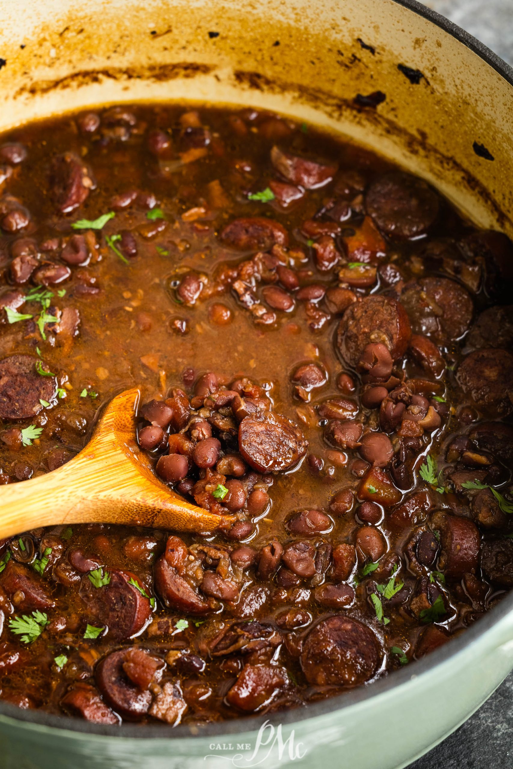 A pot of cooked red beans and sausage in a thick brown sauce, being stirred with a wooden spoon. Some green herbs are sprinkled on top.